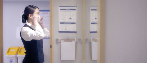 A person in a white shirt and dark vest standing in a room with a mirror, a door, and informational posters on the wall.