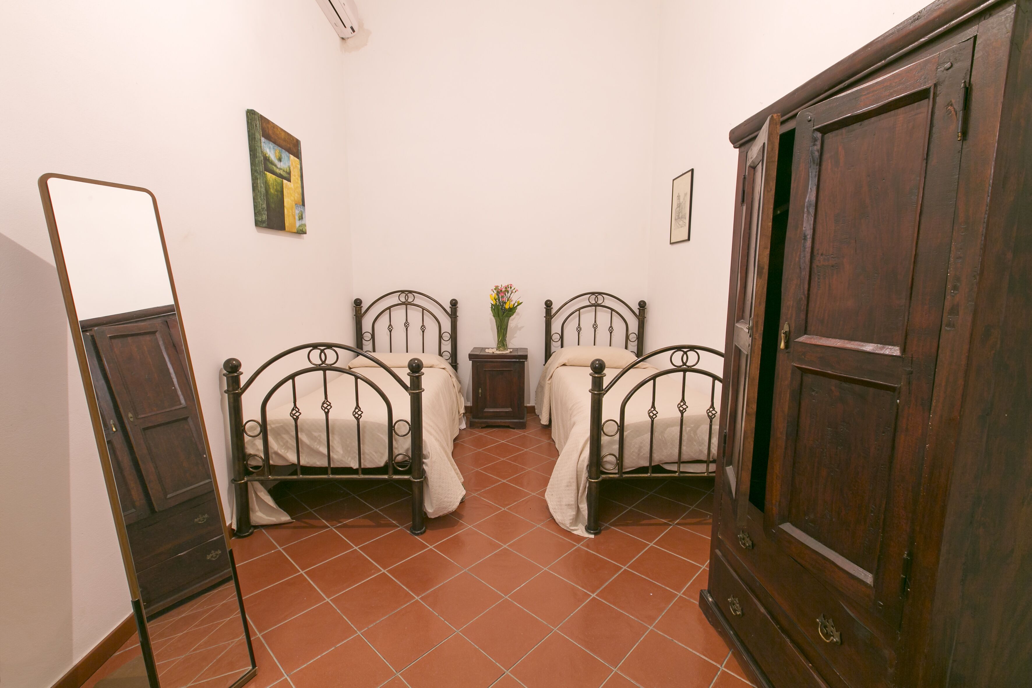 Una camera da letto con due letti in metallo, un'armadietto in legno, un comodino con un vaso e uno specchio appeso al muro.