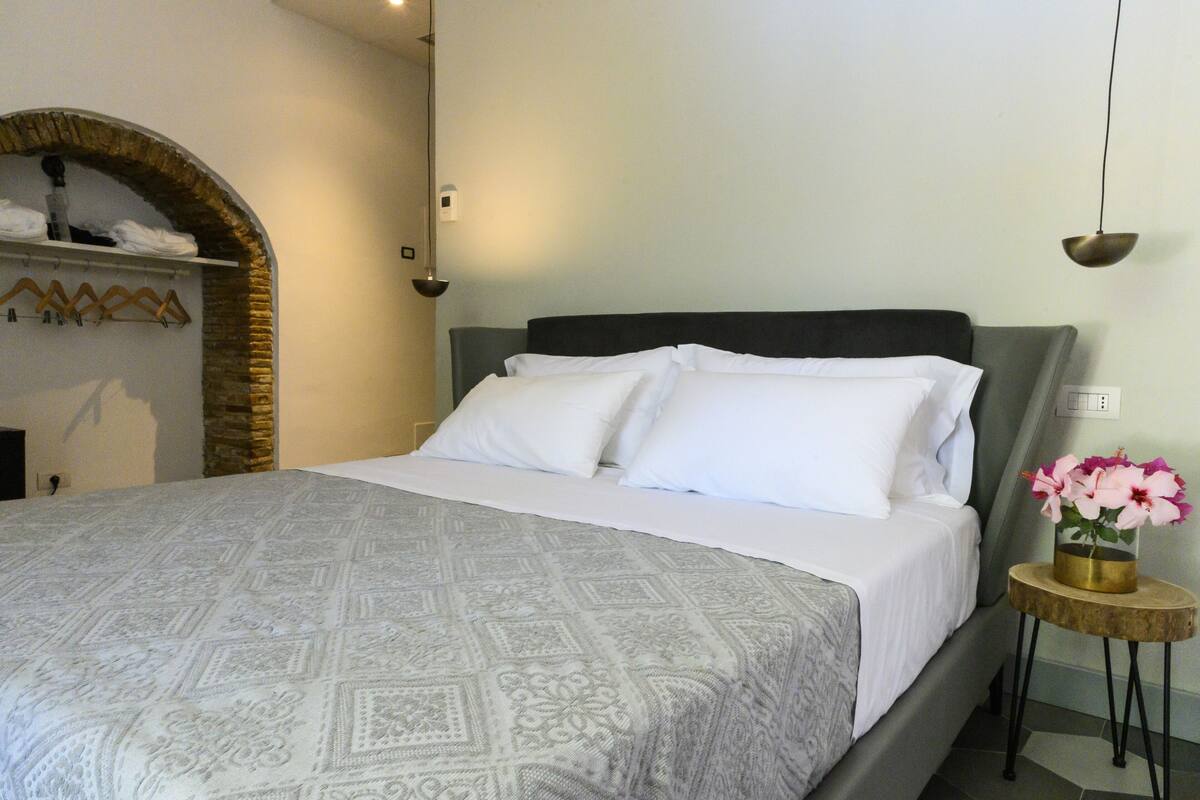 A neatly made bed with a patterned bedspread, a small bedside table with a vase of flowers, and a wall-mounted shelf with folded towels.