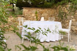 A round outdoor table set for four with white chairs, a stone wall backdrop, and greenery in the foreground.
