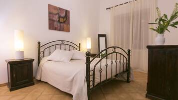 A bedroom with a metal bed frame, a bedside table with a lamp, a nightstand, a mirror, a dresser, a painting on the wall, and a window with curtains.