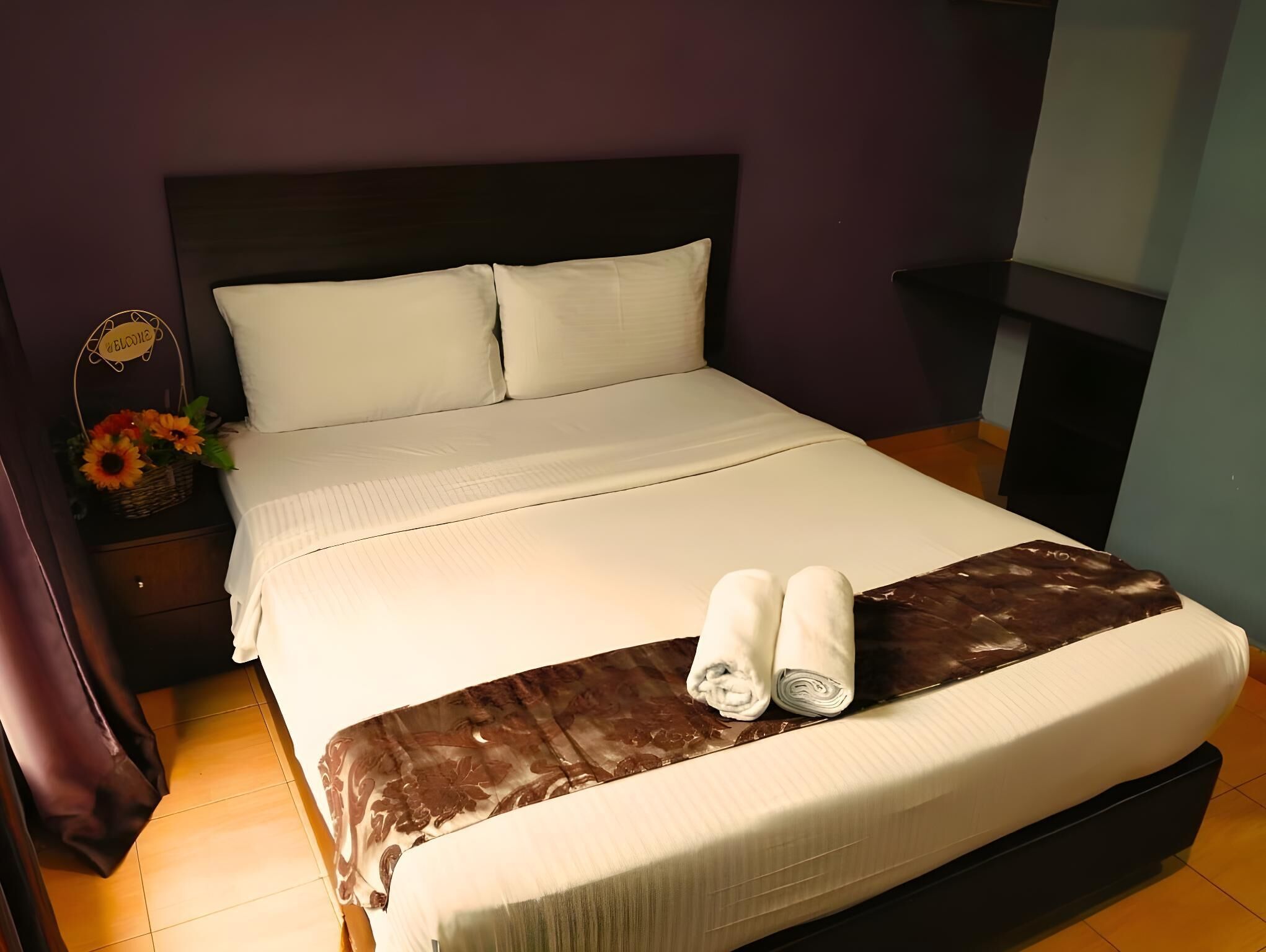 A neatly made bed with white linens, a brown throw blanket, and two rolled towels on the bed.
