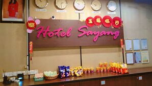 A hotel reception desk with clocks indicating different time zones, a large sign reading "Hotel Sayang," and various snacks and amenities on the counter. - Hotel Sayang (Kulai)