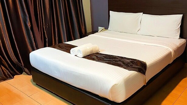 A neatly made bed with white linens and a brown accent, a dark brown curtain, and a wall-mounted sign with a blue background and white text. - Hotel Sayang (Kulai)
