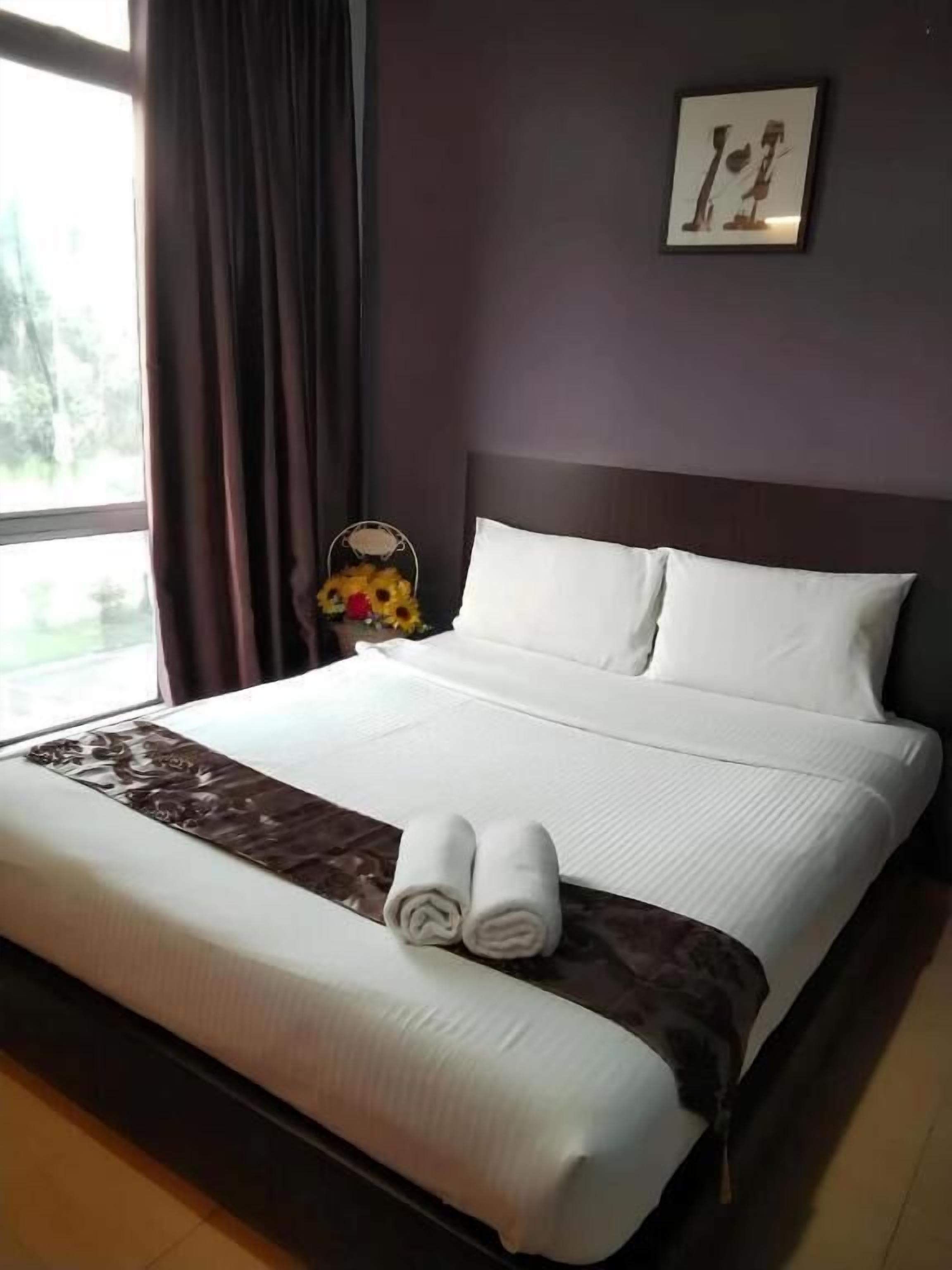 A neatly made bed with white linens and two rolled towels, a dark brown headboard, and a framed picture on the wall.