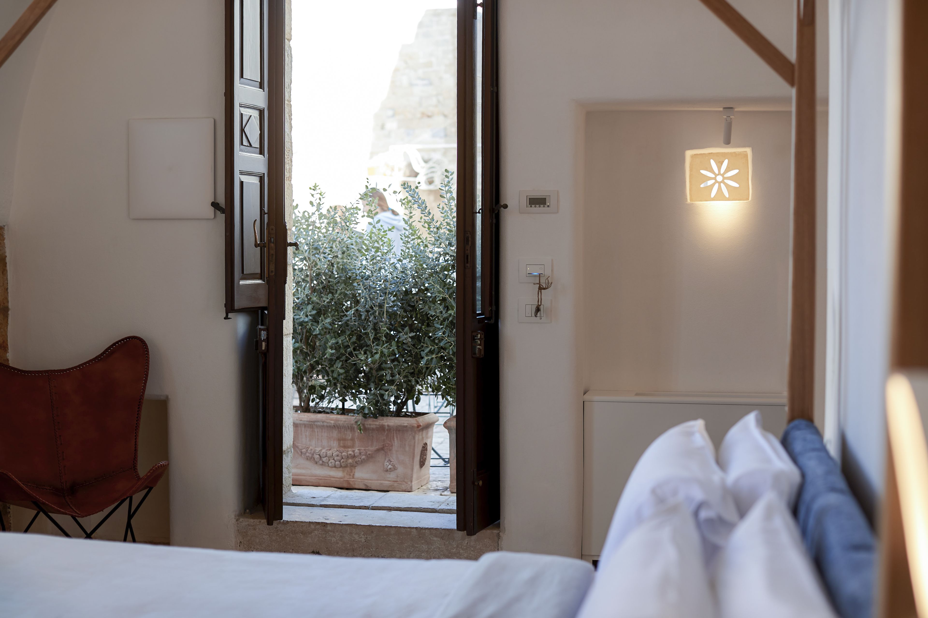 A bedroom with a bed, a chair, a potted plant, and a view of the outside.