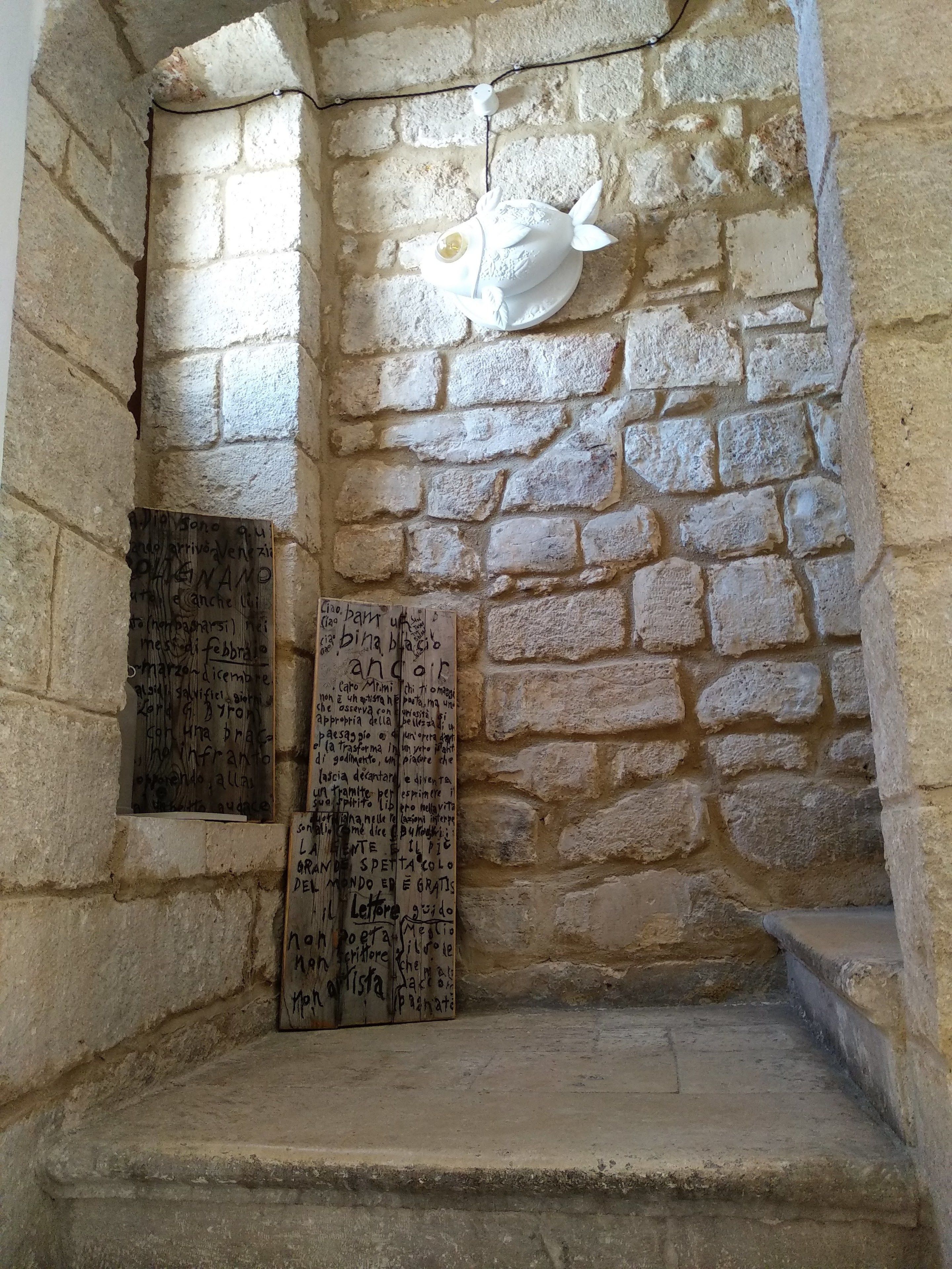 Habitación de paredes de piedra con una pequeña escalera, un aplique blanco fijado a la pared y dos hojas de papel con texto manuscrito.