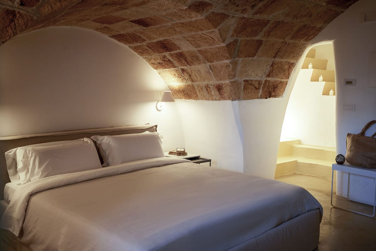 Un dormitorio con techo de piedra, una cama con ropa blanca, una mesita de noche y una lámpara.