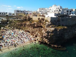 Uma praia cheia, com águas cristalinas, falésias rochosas e edifícios brancos ao fundo.