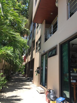 A narrow street with buildings on either side, a visible storefront with large glass windows, and a staircase leading to an upper level. - Smile Home (Patong)