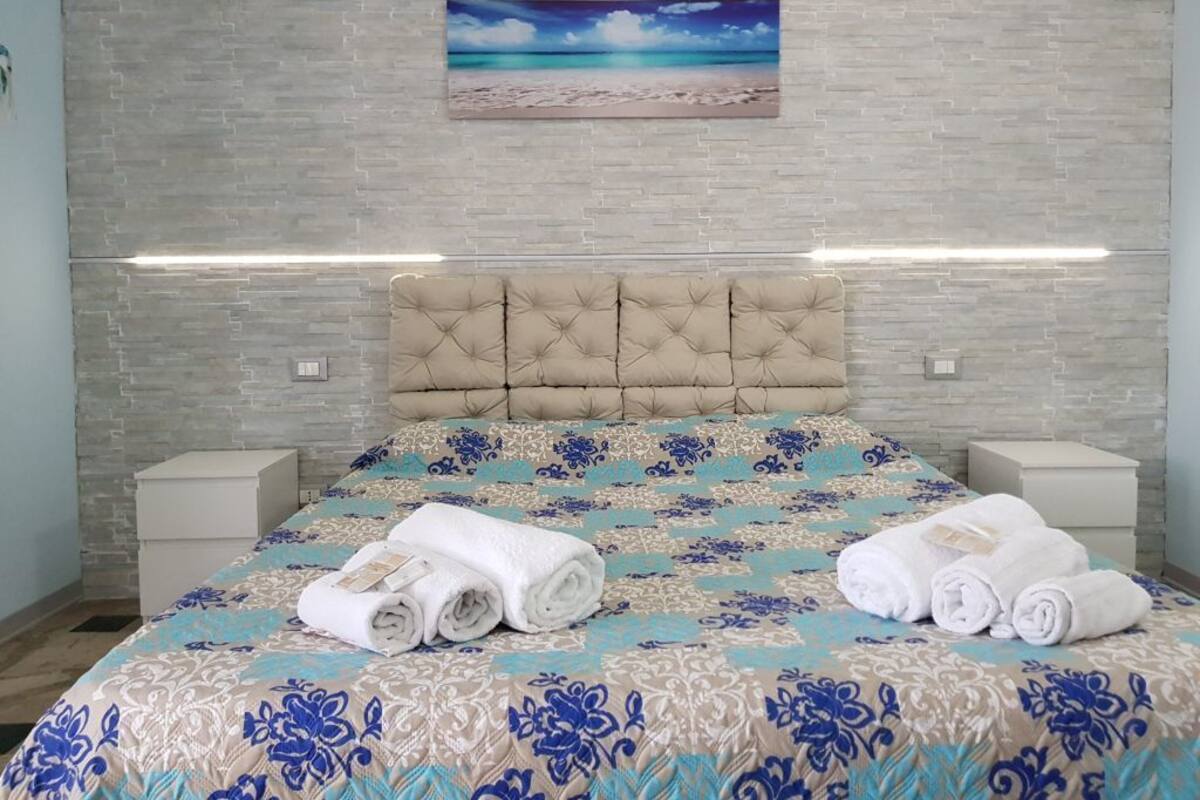 A hotel room with a bed, headboard, nightstands, and a painting of a beach scene.