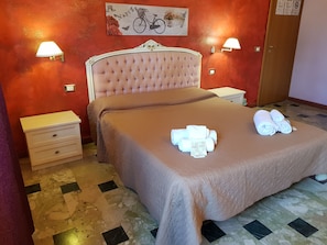 A neatly made bed with a beige bedspread and two rolled towels on the bed. There is a nightstand with a lamp and a small table beside the bed. The room has a red wall with a nature-themed artwork above the bed. - B&B Catania Mare (Catania)