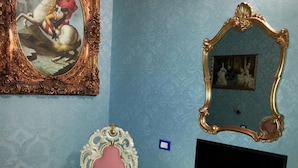 A room with a blue patterned wall, a white dresser, a flat-screen TV, a pink upholstered chair, and two ornate mirrors. - B&B Catania Mare (Catania)