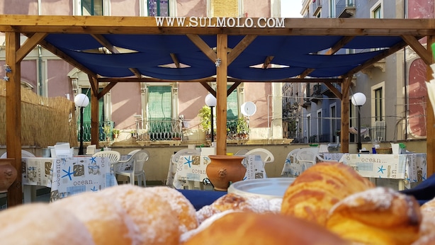 An outdoor café with tables covered by blue umbrellas, a wooden pergola, and a view of a building with balconies. - B&B Catania Mare (Catania)