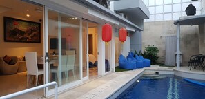 Un área interior de piscina moderna con pared de vidrio, sillas tipo bean bag azules y una zona de comedor con mesa y sillas.