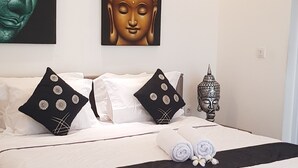 A neatly made bed with white bedding and decorative pillows, a wall-mounted Buddha statue, and a small decorative lantern. - Villa Fei (Jimbaran)