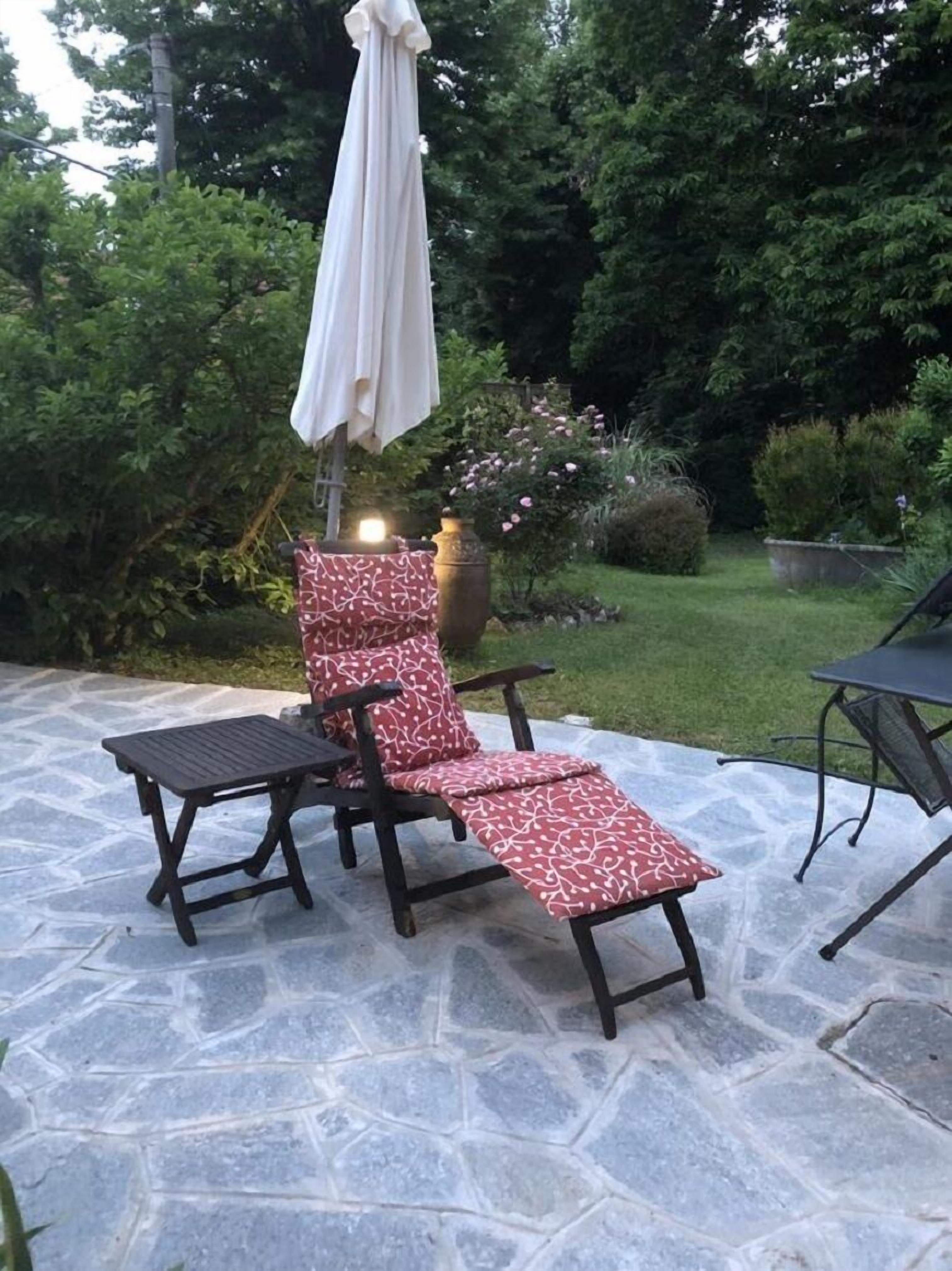 Un patio con suelo de piedra, una silla de lounge de madera con un cojín estampado y una sombrilla blanca.