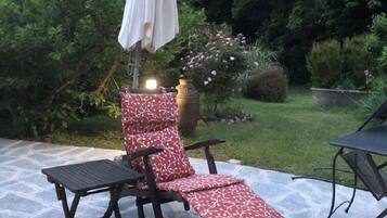 Un patio con suelo de piedra, una silla de lounge de madera con un cojín estampado y una sombrilla blanca.
