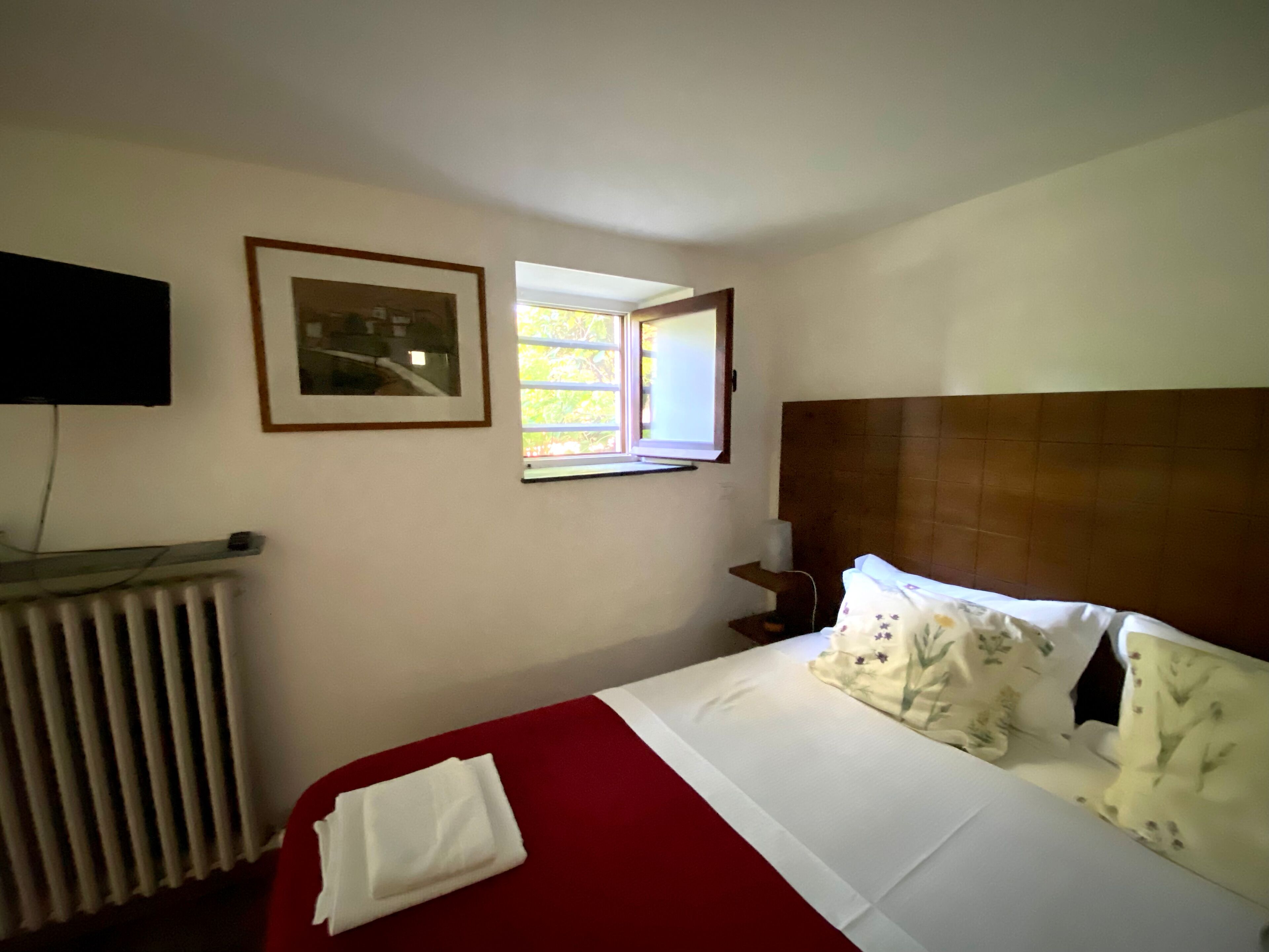 A hotel room with a bed, a window, a radiator, and a TV mounted on the wall.