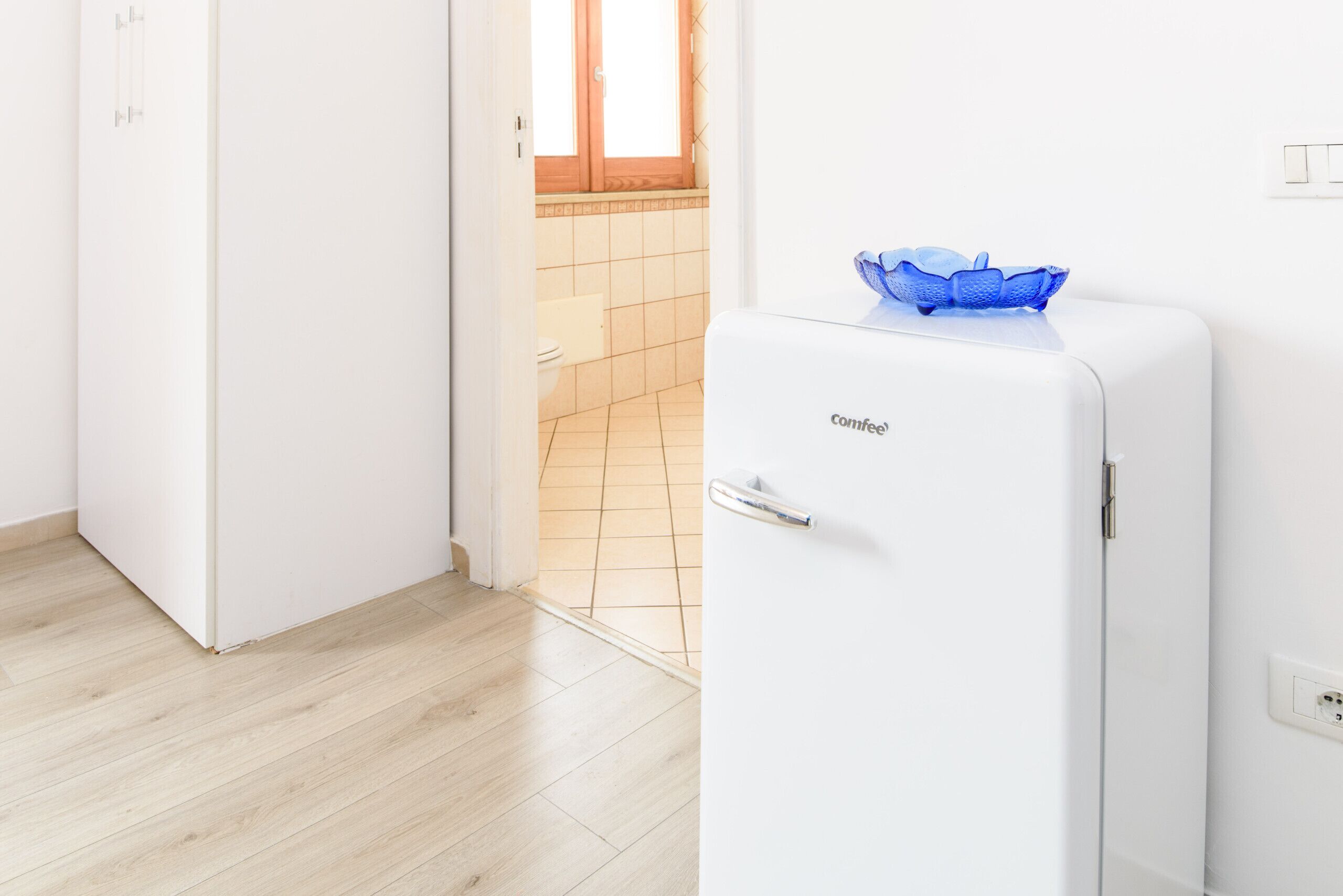 A compact kitchen with a white refrigerator, a blue glass bowl on top, and a wooden window sill.