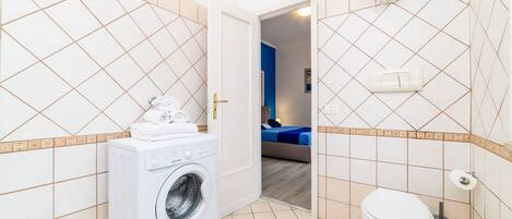A compact bathroom with a washing machine, toilet, and a visible bedroom in the background.