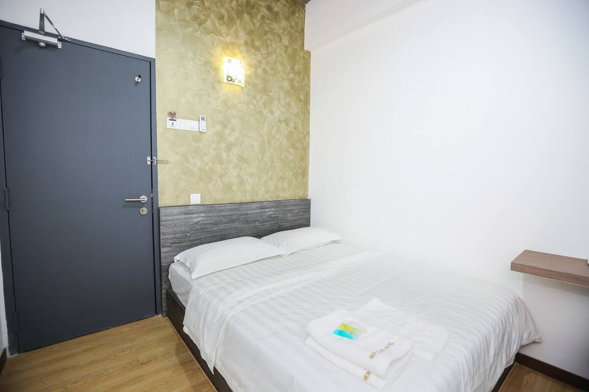 A hotel room with a bed, a wooden floor, a grey door, a textured wall, and a small shelf.