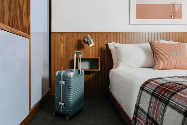 A hotel room with a bed, a wooden headboard, a wall-mounted lamp, a suitcase, and a framed picture on the wall.