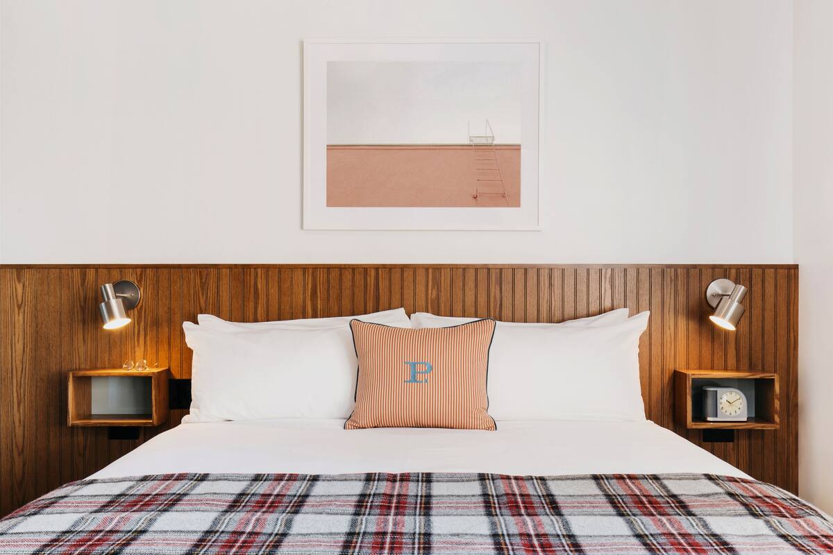 Hotellihuone, jossa on puinen pääty, ruudullinen peitto sängyllä, kehystetty kuva seinällä sekä yöpöydät, joissa on lamput ja kellot.