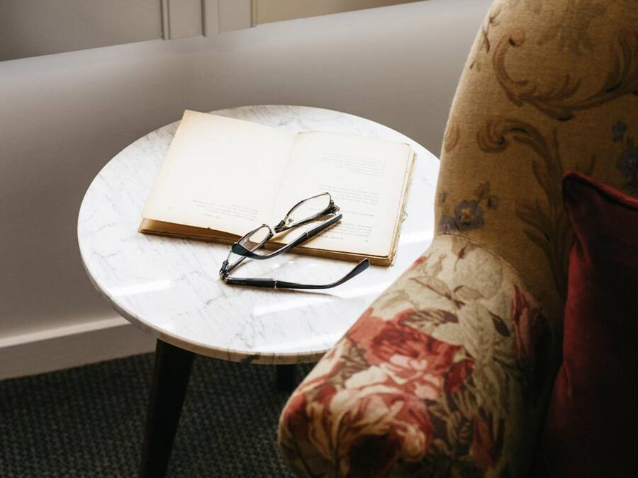 Een kleine ronde tafel met een boek en brillen, een bloemenbank en een raam met jaloezieën.