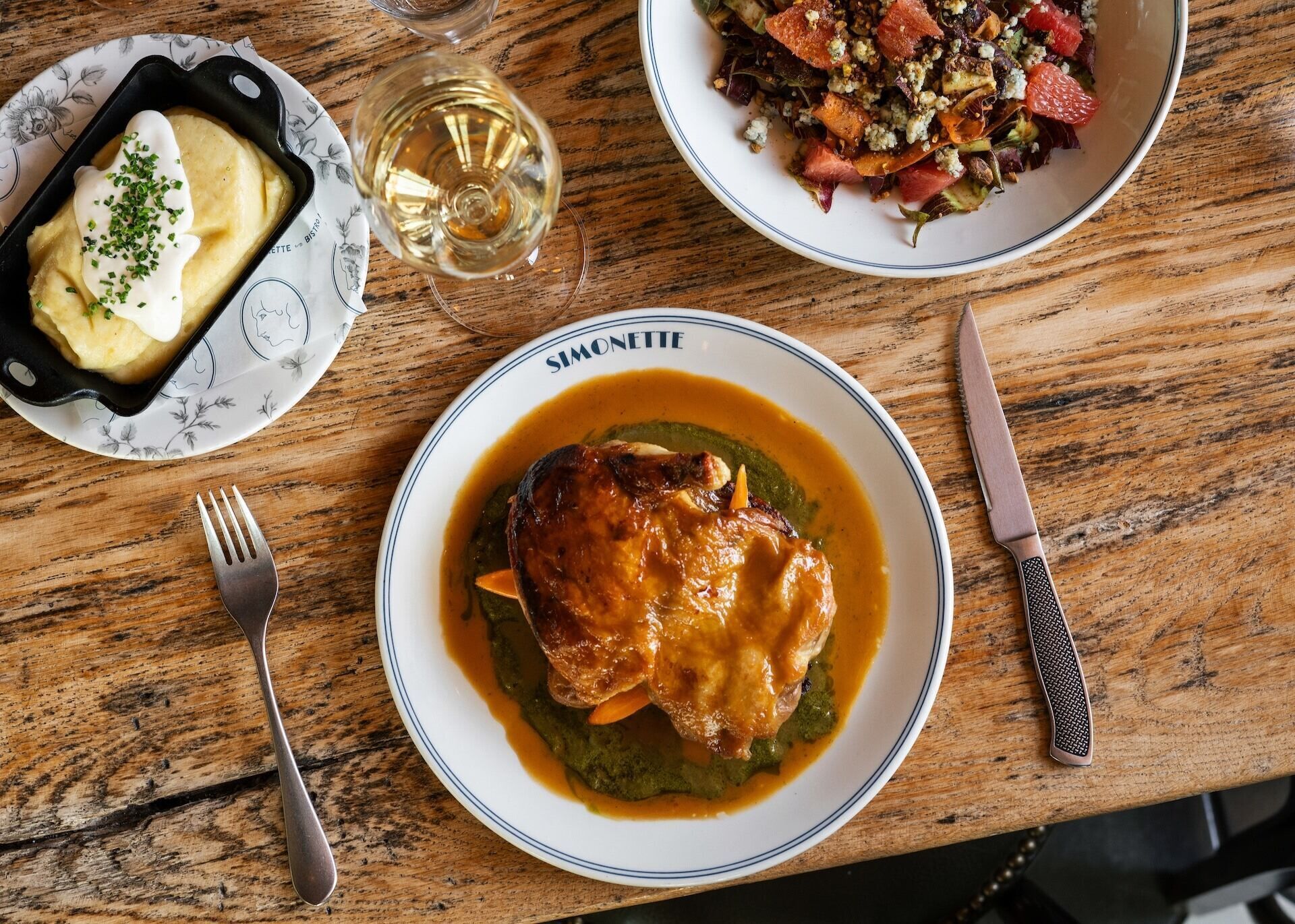 A wooden table with a plate of food, a fork, a knife, and a glass of white wine.