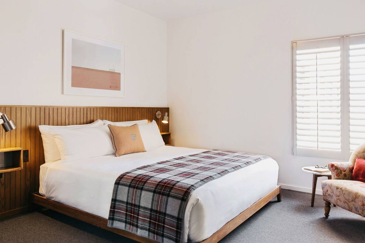 Hotellihuone, jossa on puinen pääty, valkoisilla lakanoiden peitetty sänky, ruudullinen heittopeitto, pieni yöpöytä lampun kanssa ja ikkuna, jossa on sälekaihtimet.