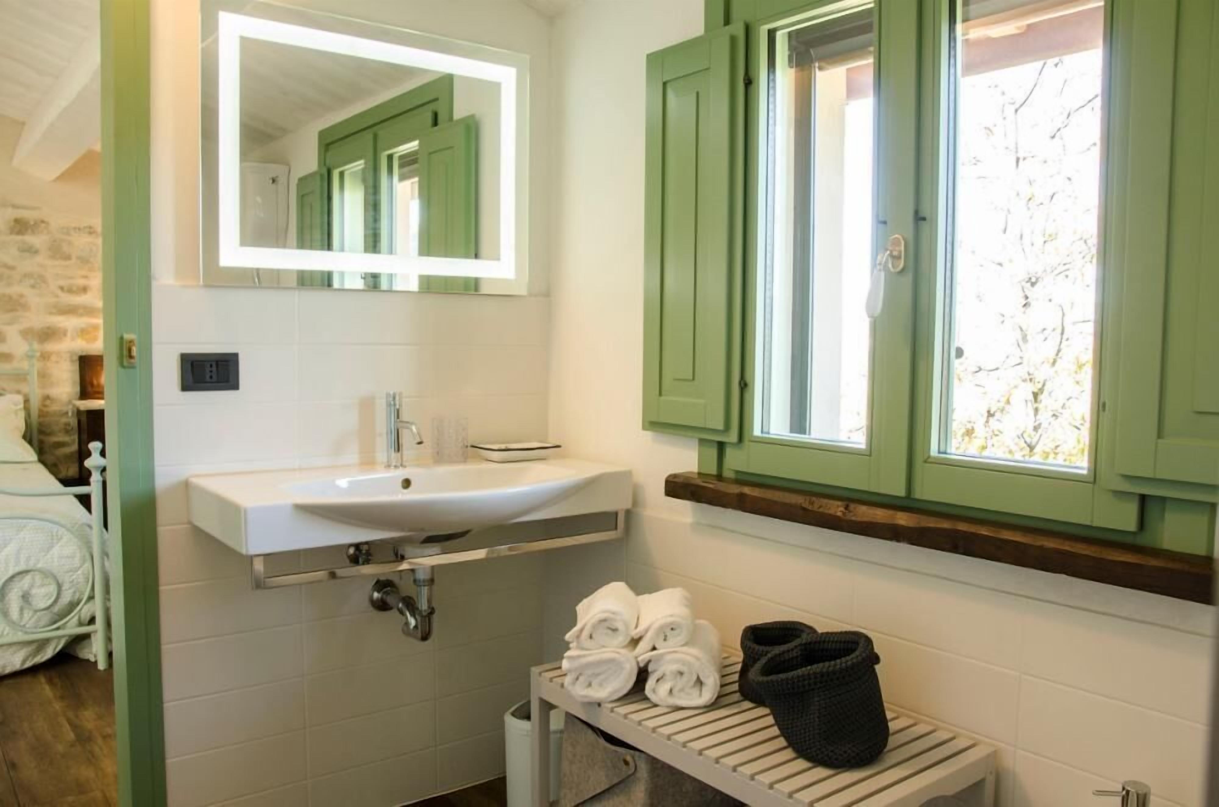 Un baño con lavamanos blanco, armarios verdes, un espejo y una ventana con contraventanas.