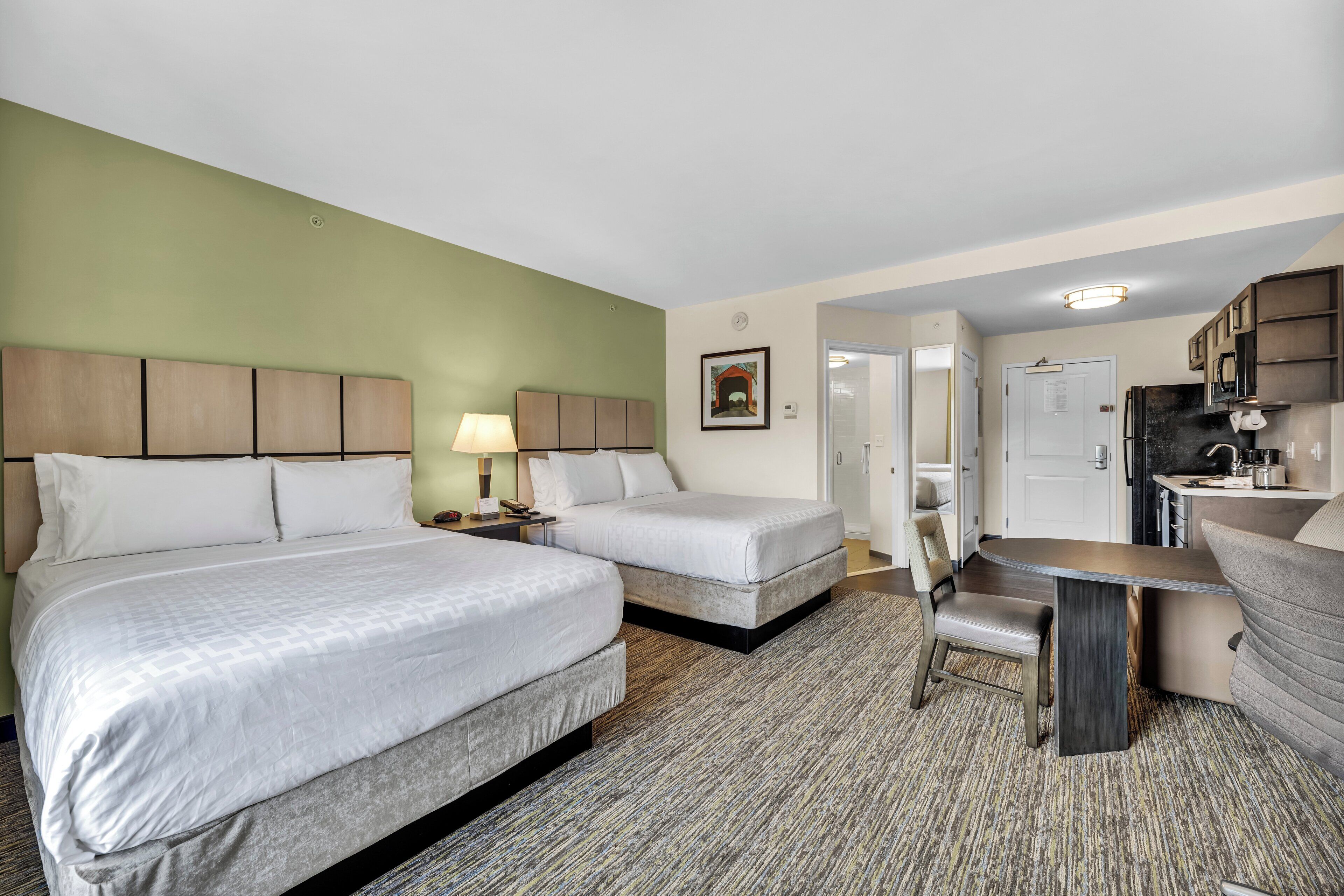 A hotel room with two beds, a desk, a chair, a kitchenette, and a door leading to another room.