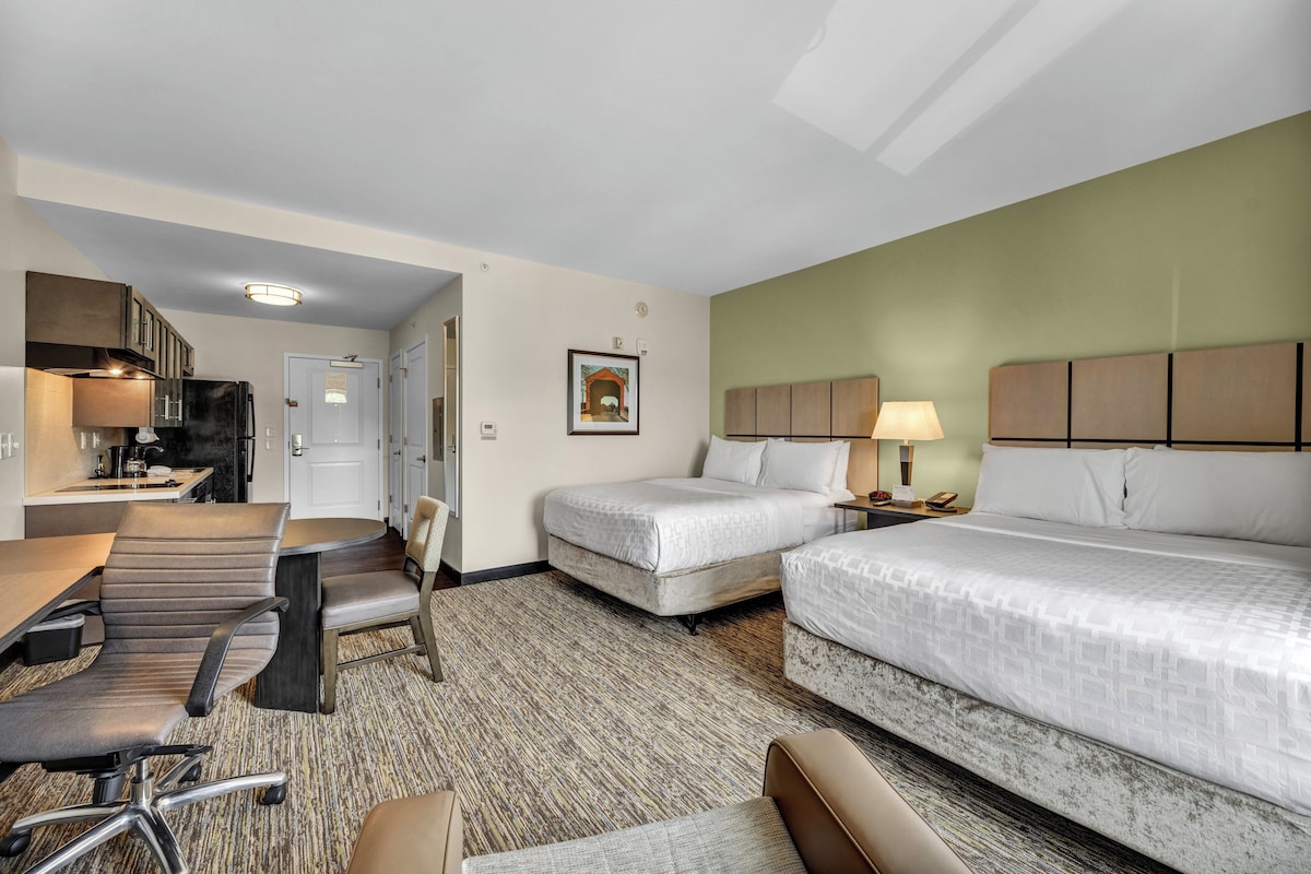Ein Hotelzimmer mit zwei Betten, einem Schreibtisch mit Stühlen, einer Küchenzeile und einem gerahmten Bild an der Wand.