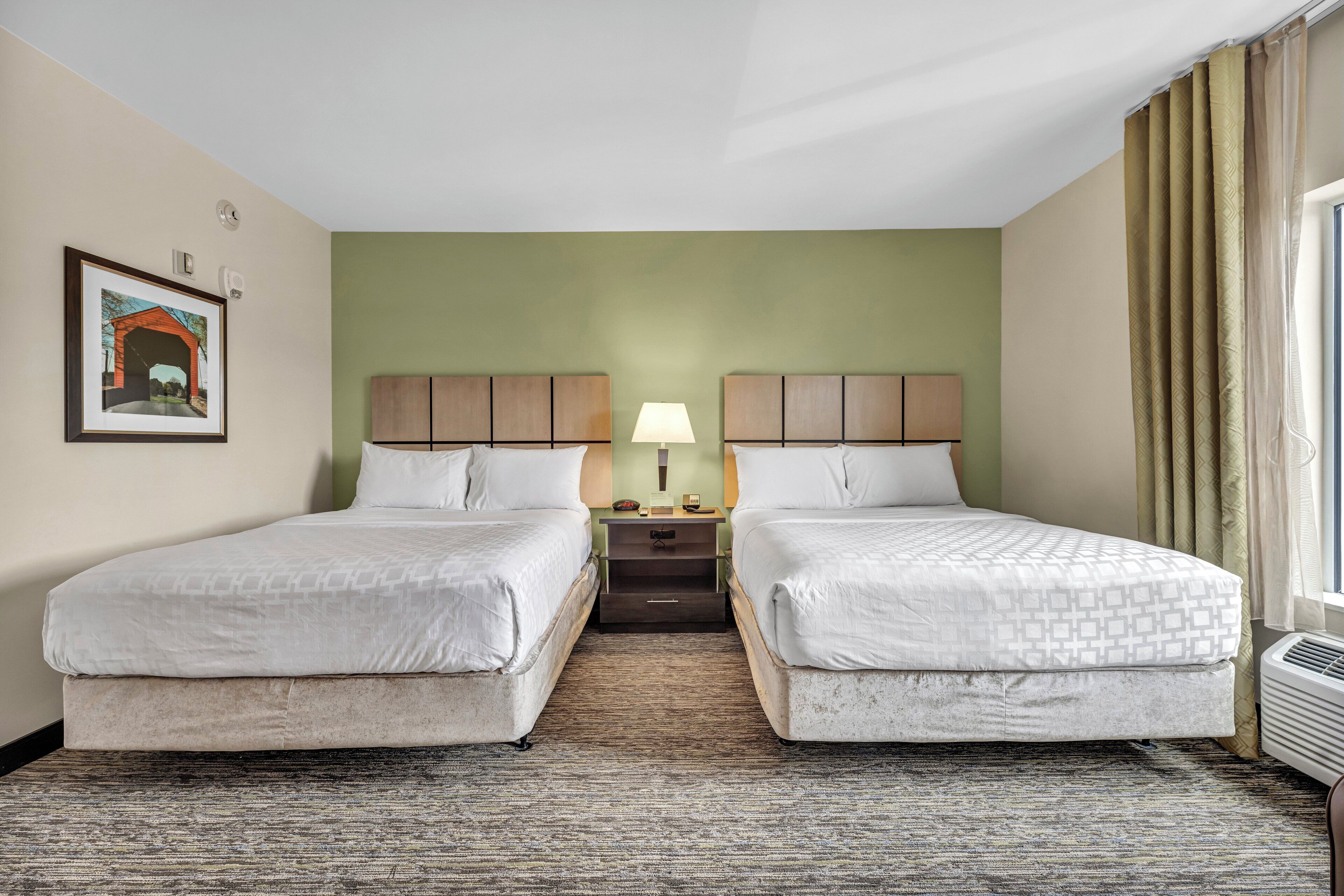 A hotel room with two beds, a nightstand with a lamp, a framed picture on the wall, and a window with curtains.