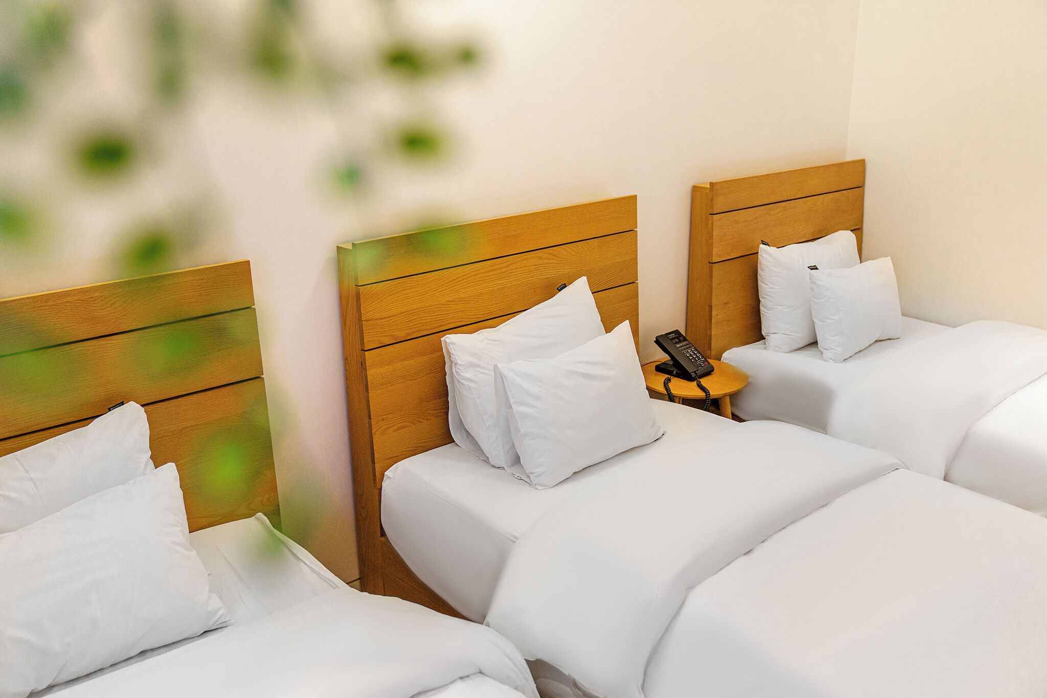 A hotel room with three beds, each with white bedding and wooden headboards. There is a small bedside table with a phone on it.