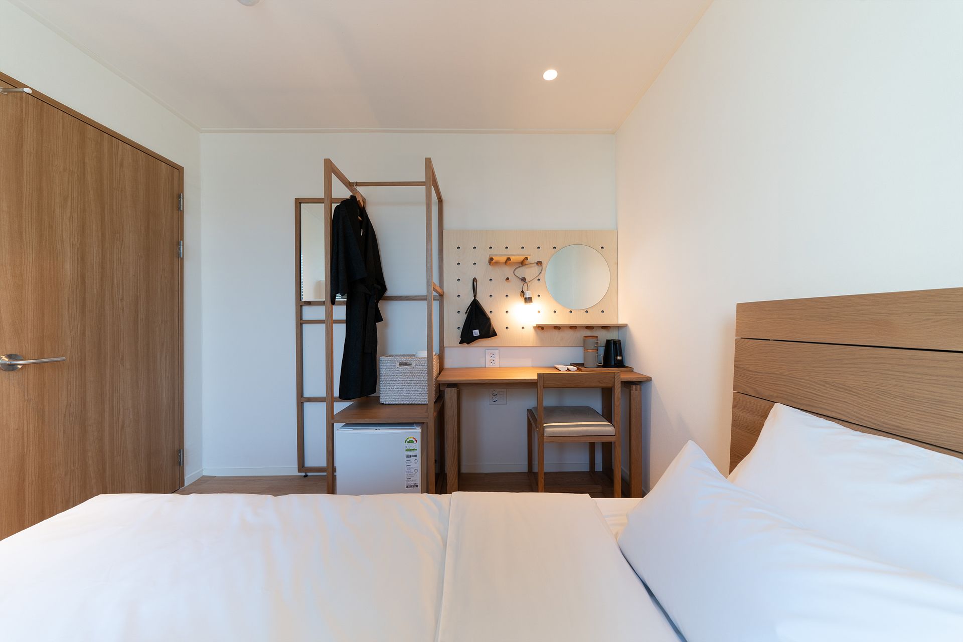 A bedroom with a bed, a desk, a chair, a wardrobe, and a wall-mounted shelf with a mirror and a small table.
