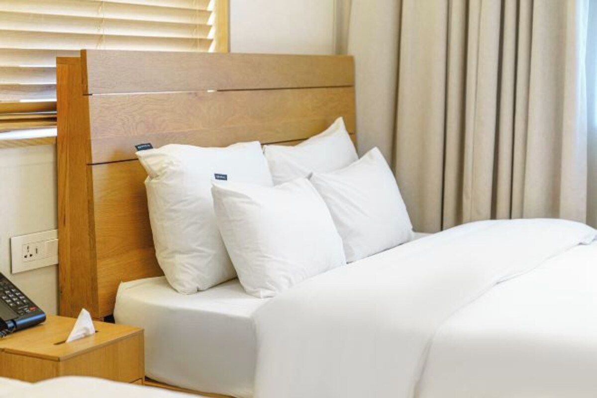 Une chambre d’hôtel avec une tête de lit en bois, du linge de lit blanc, une table de chevet avec un téléphone et des mouchoirs, et une fenêtre avec des stores.