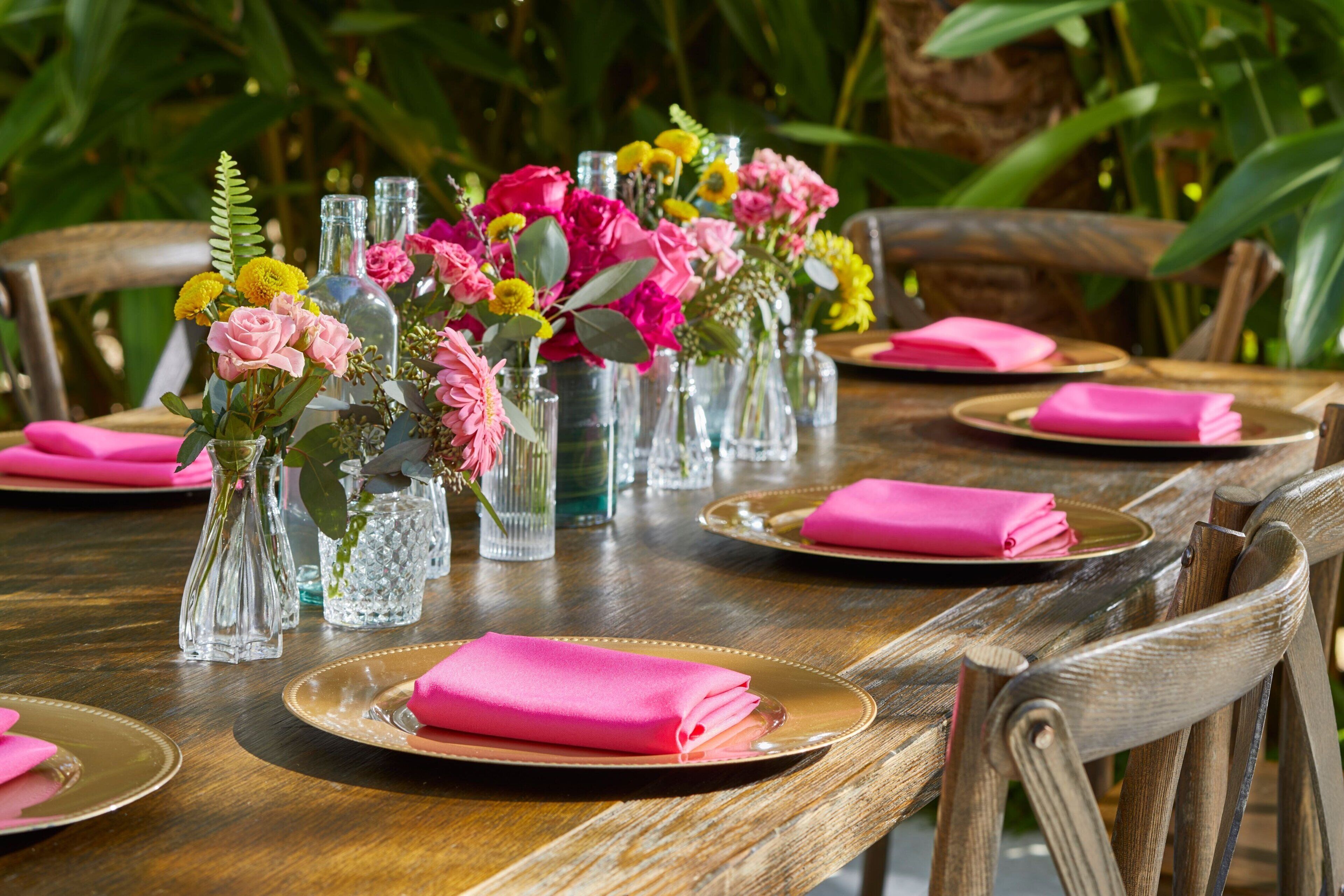 Een houten eettafel gedekt met roze servetten, glazen vazen met bloemen en gouden borden.