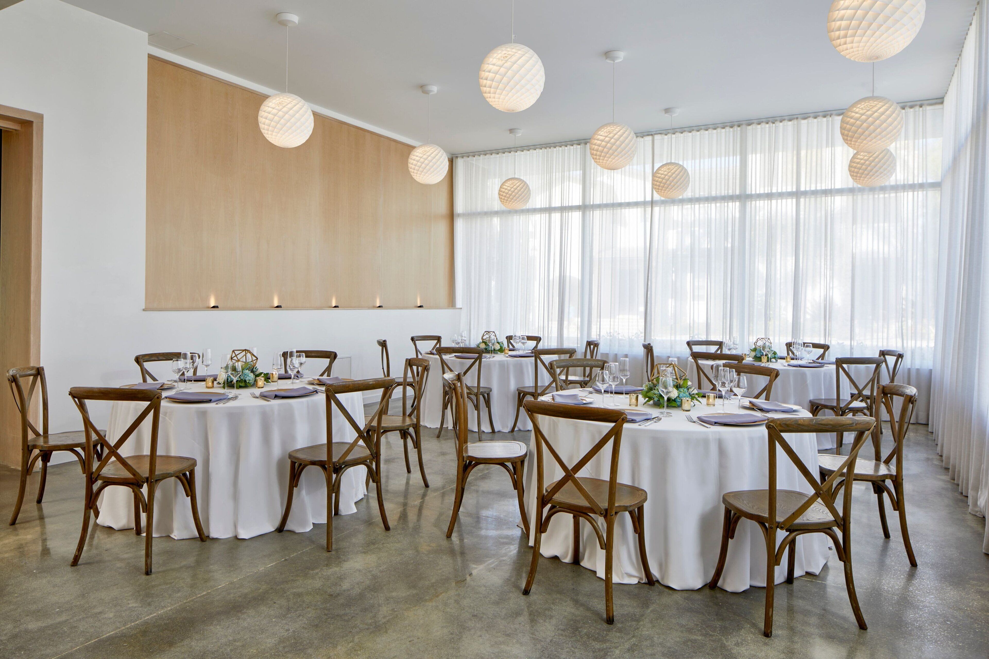 A spacious room with multiple round tables set for dining, wooden chairs, and pendant lighting.