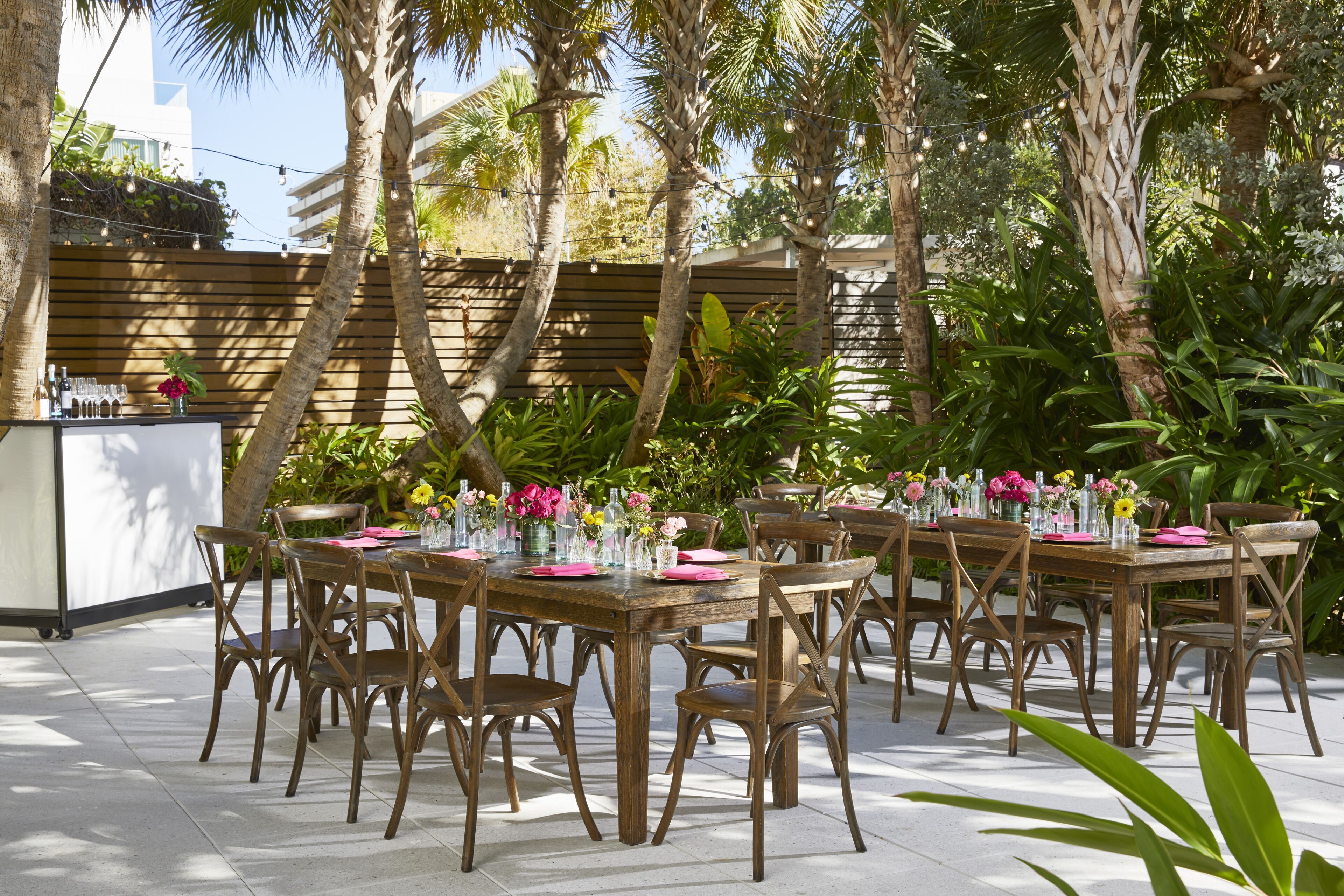 An outdoor dining area with wooden tables and chairs, surrounded by lush greenery and palm trees.