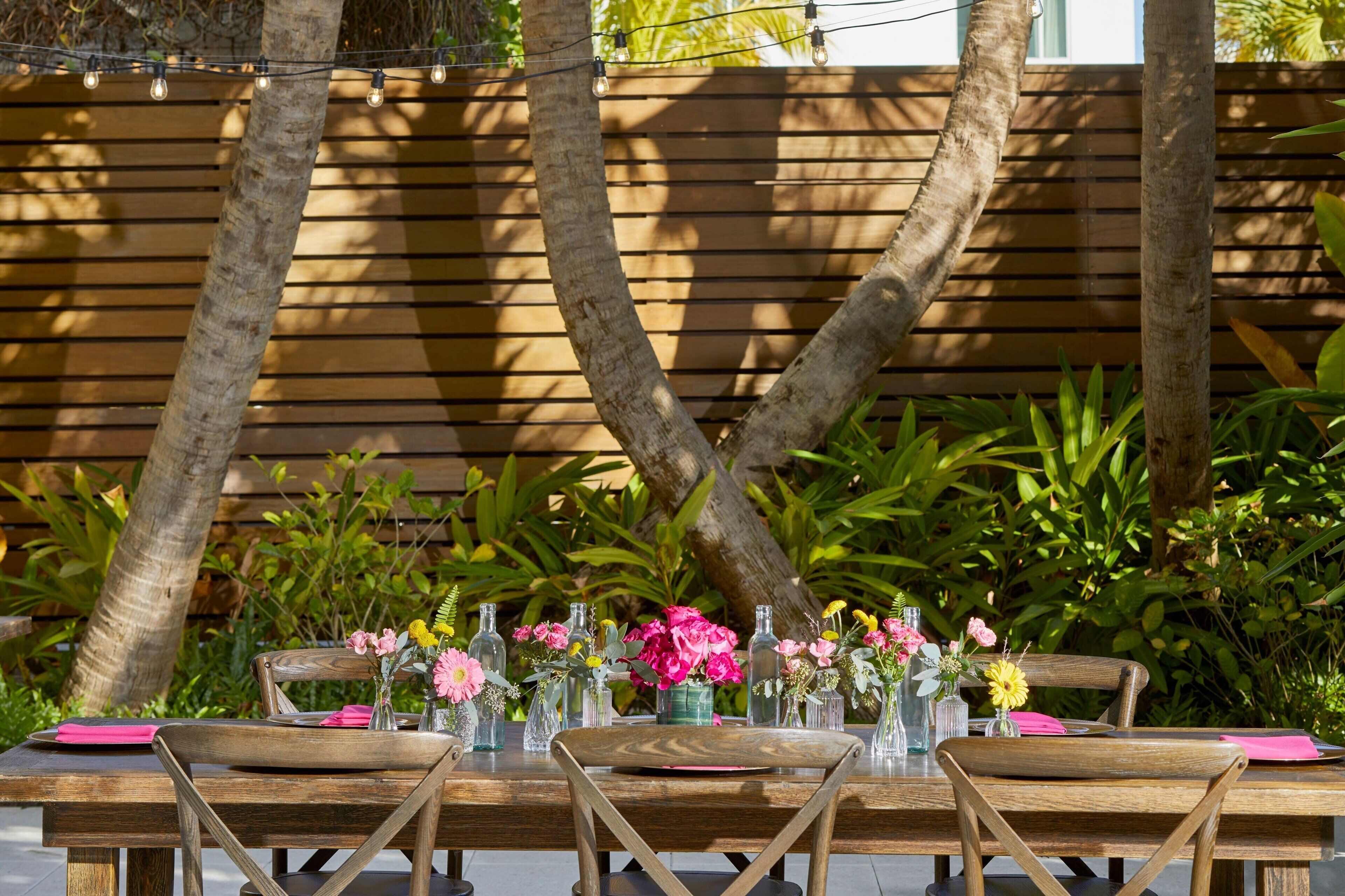 An outdoor dining area with wooden tables and chairs, surrounded by lush greenery and palm trees.