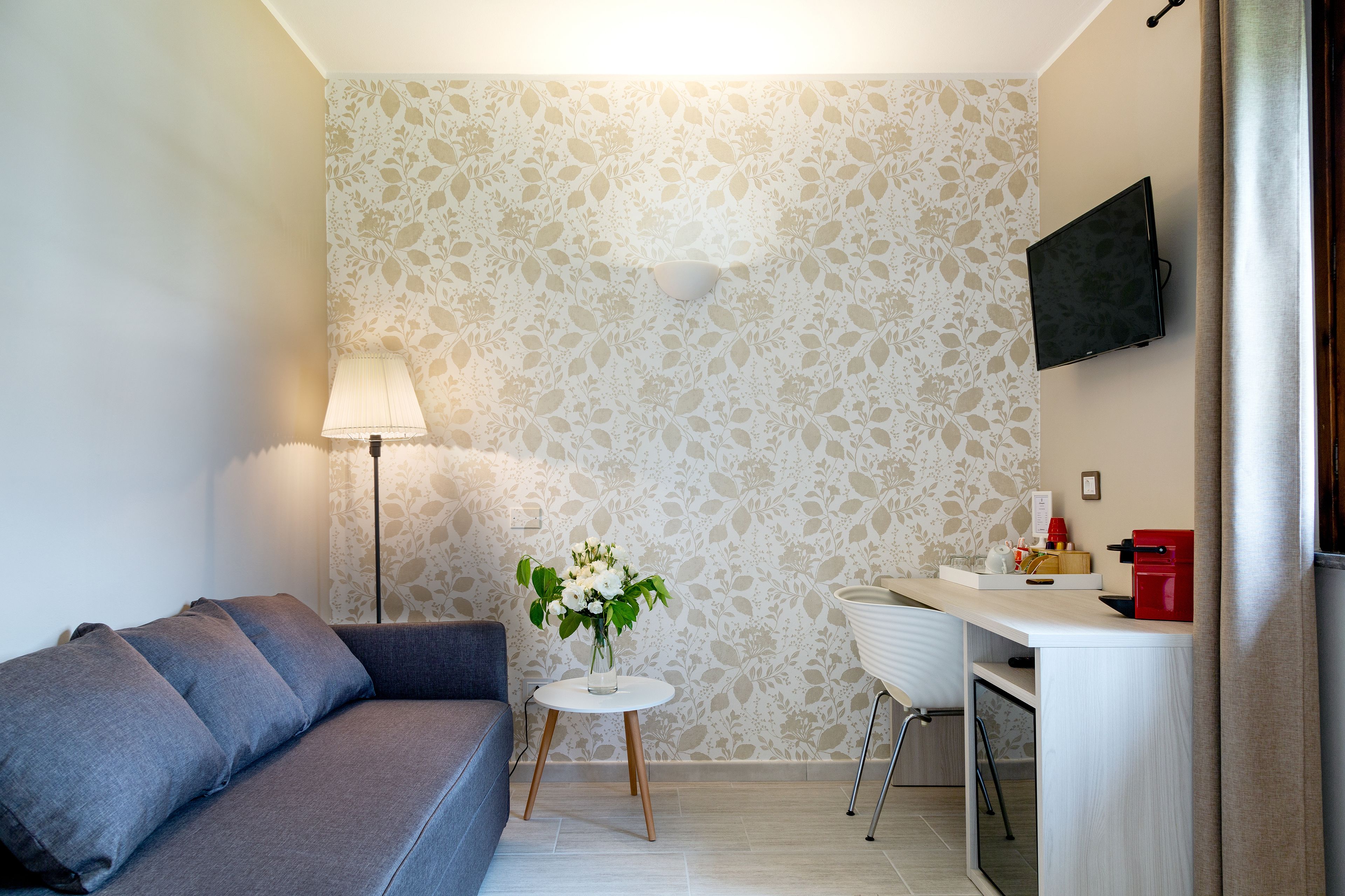 A modern living room with a grey sofa, a small round table with a vase of flowers, a lamp, a TV mounted on the wall, and a kitchenette area with a red appliance.