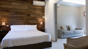 A bedroom with a wooden headboard, a bed with a patterned bedspread, a nightstand with a lamp, a grey sofa, and a wicker chair. - Tenuta Lamborghini Golf & Resort (Panicale)