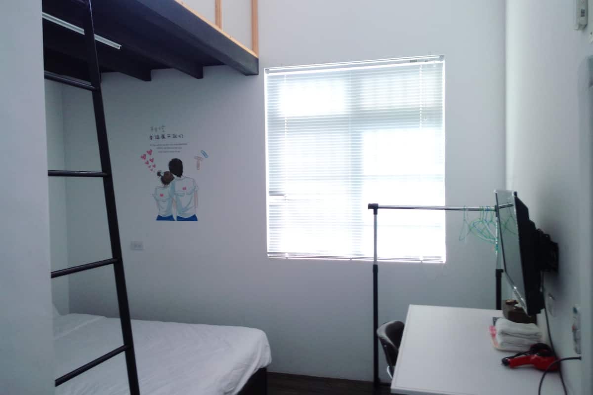 A dormitory room with bunk beds, a desk, a chair, a window with blinds, and a poster on the wall.