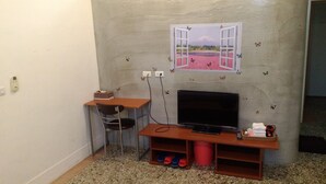 Habitació petita amb televisió sobre un suport de fusta, una cadira, una taula petita i una estant mural amb una cistella.