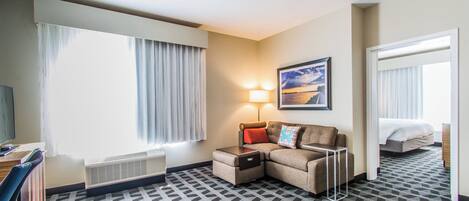 A hotel room with a sectional sofa, a desk, a standing lamp, a framed picture on the wall, and a patterned carpet.