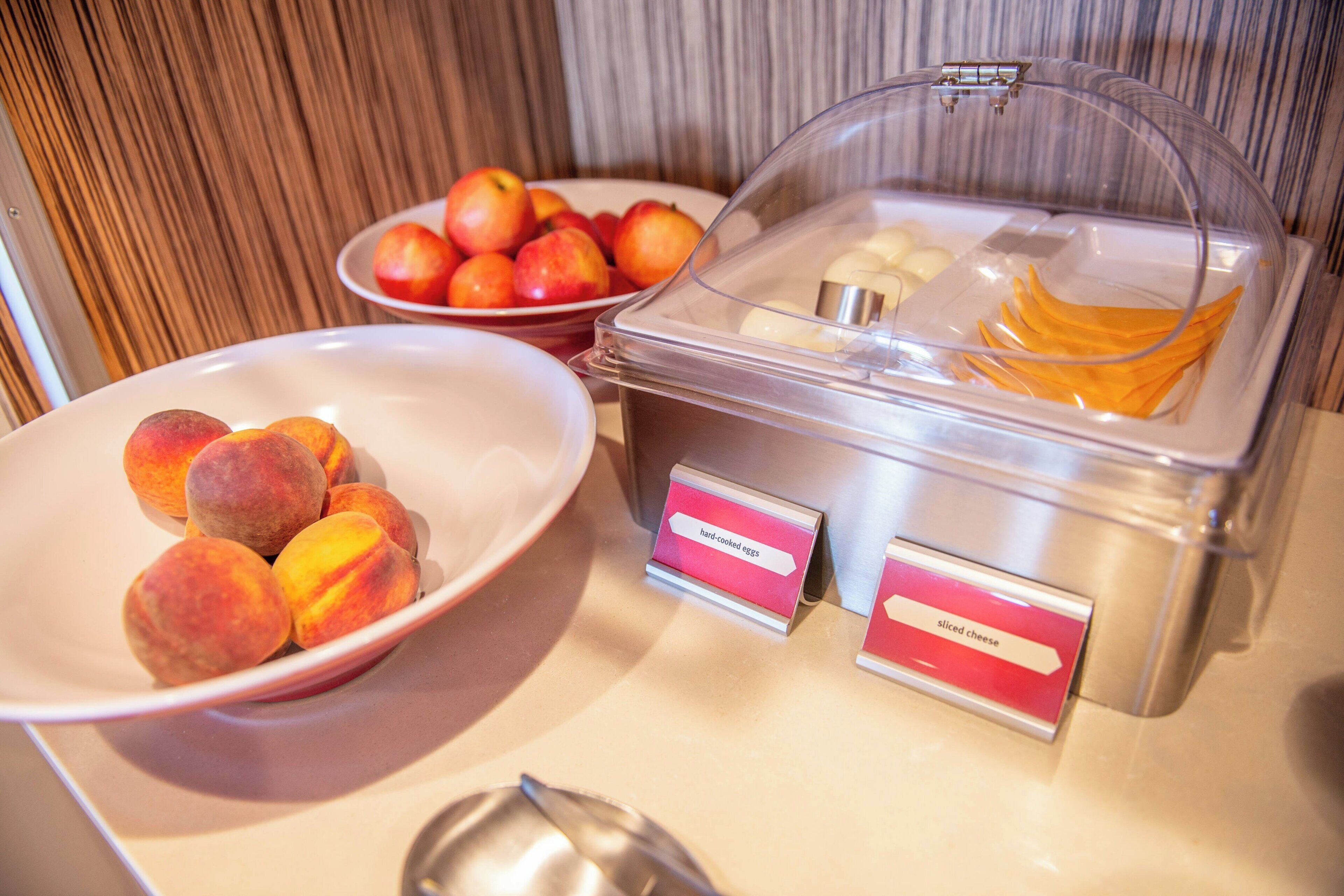 A buffet with labeled containers of hard-cooked eggs and sliced cheese, and a bowl of peaches.