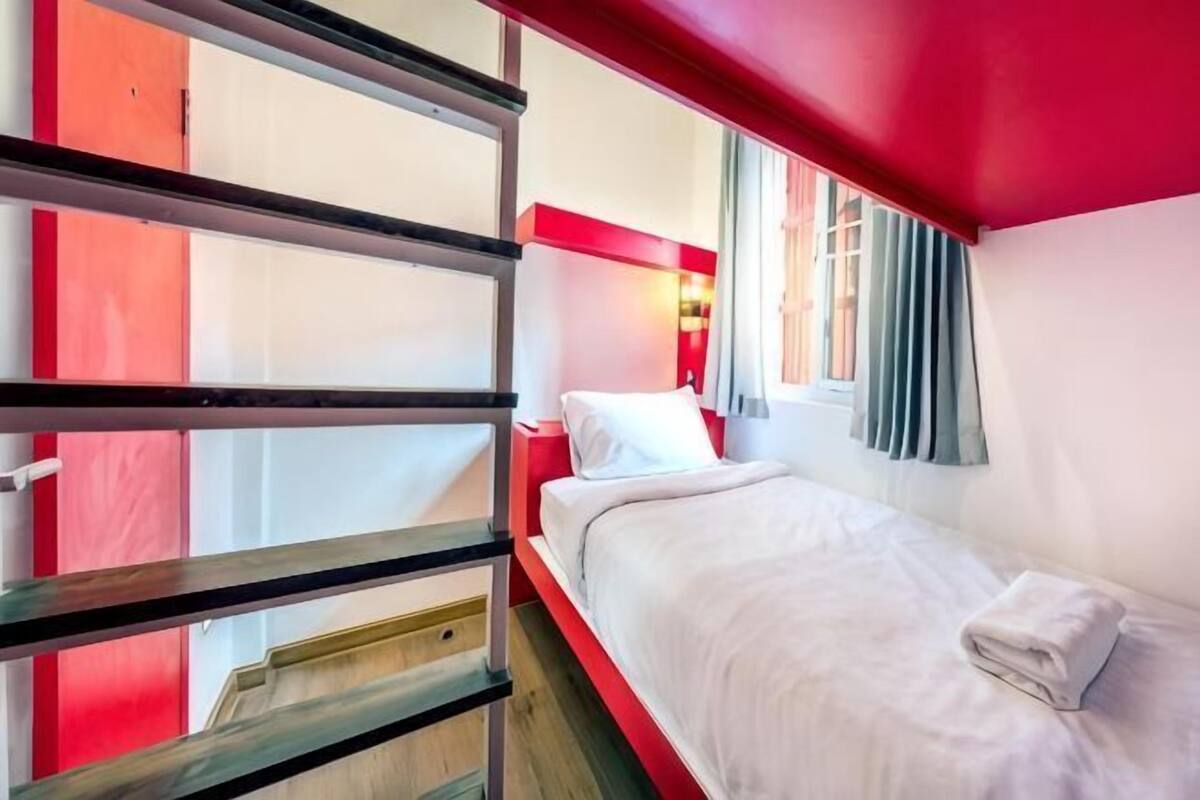Un lit simple avec une couette blanche, une tête de lit rouge et une fenêtre avec des rideaux.