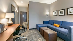 Habitación de hotel con un escritorio de madera, un sofá azul, un pequeño reposapiés, una silla, una lámpara y cuadros en la pared.